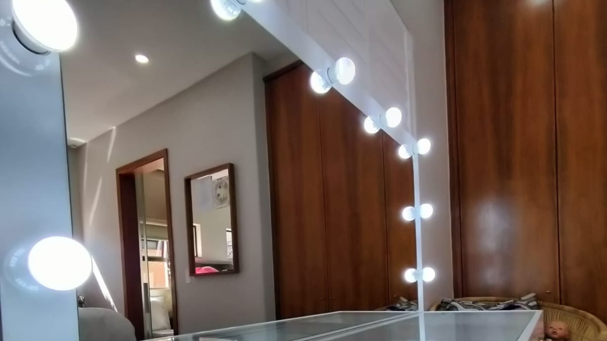 Mirror with light bulbs along the edge in a room with wooden cabinets and a framed picture on the wall.
