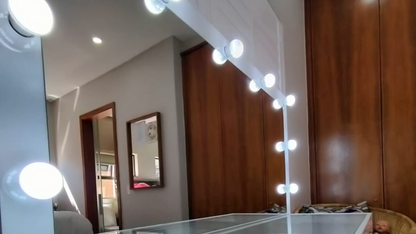 Mirror with light bulbs along the edge in a room with wooden cabinets and a framed picture on the wall.